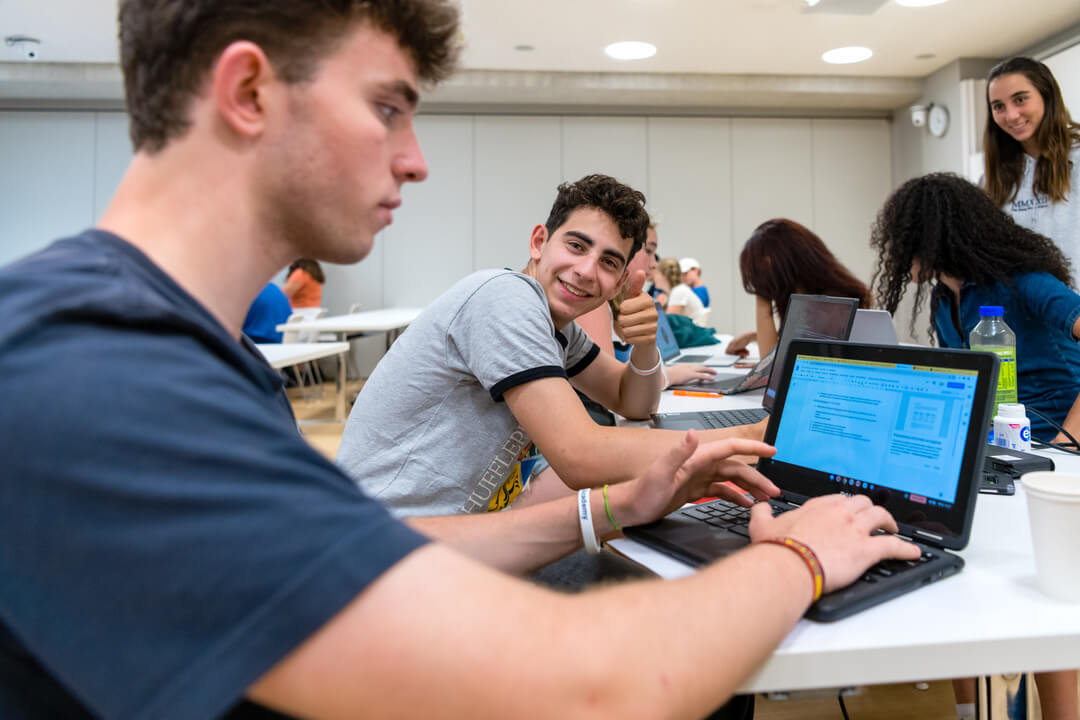 An Oxford Scholastica Academy summer school student holds a thumbs up to the camera whilst working with friends